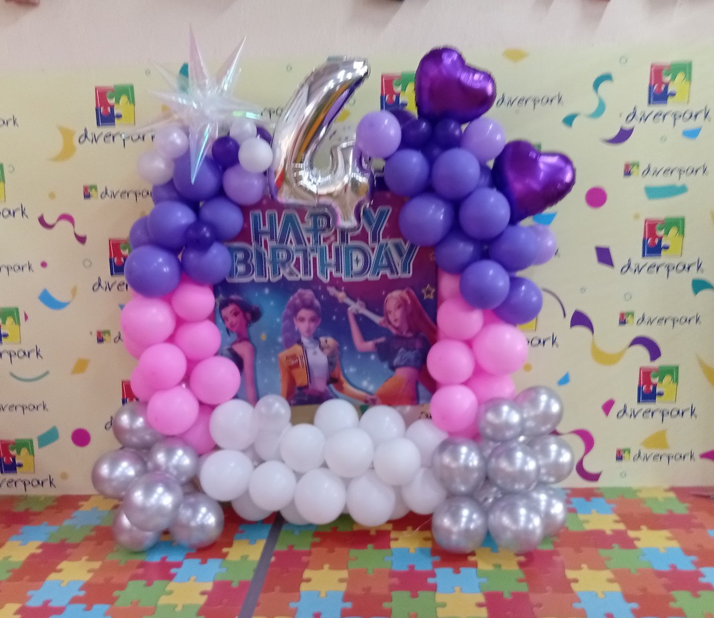 Photocall de cumpleaños con arco de globos morados, rosas y plateados sobre el suelo de puzzle de diverpark